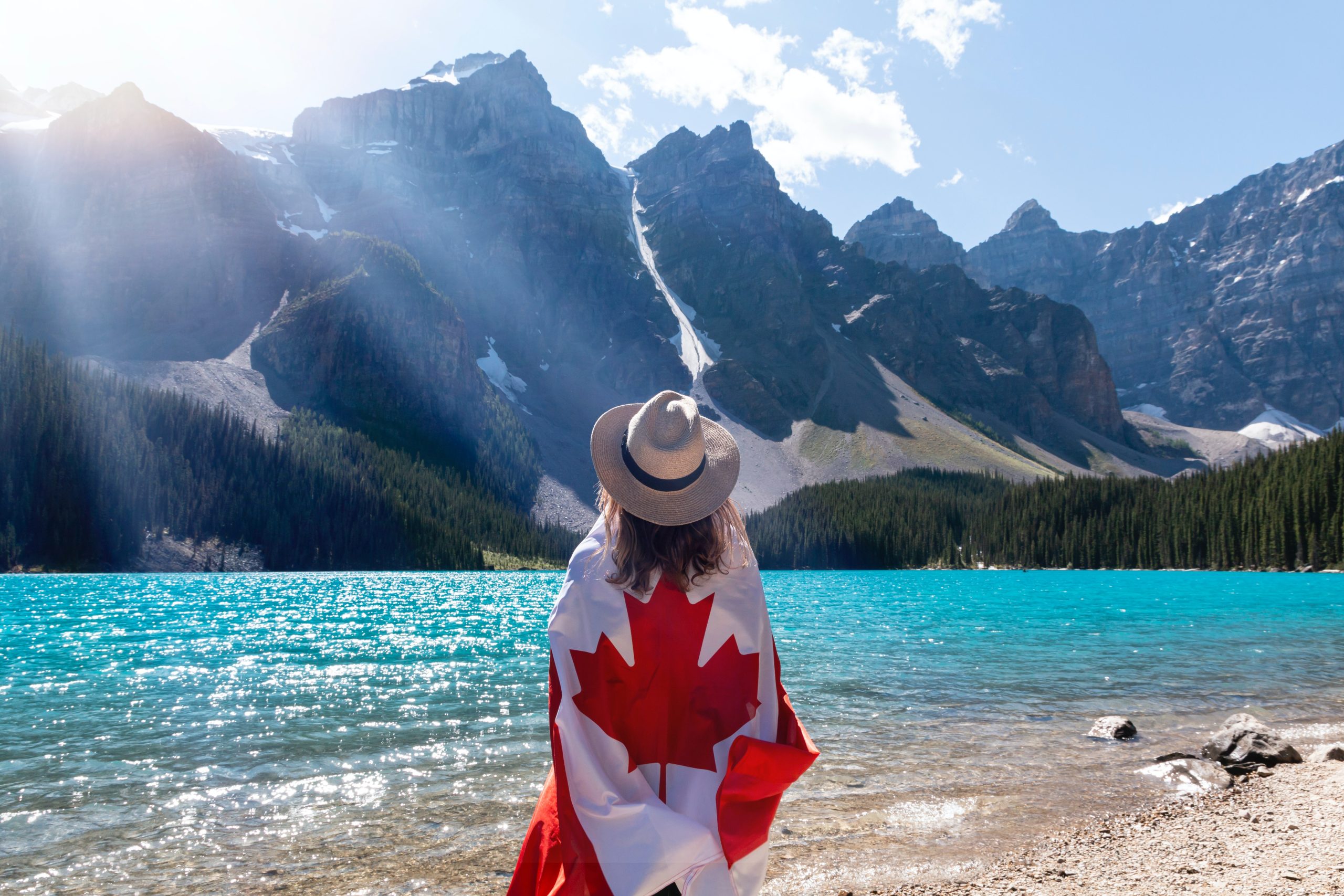 A woman wears a beige hat and is wrapped in a Canadian flag. Mountains are in the background.