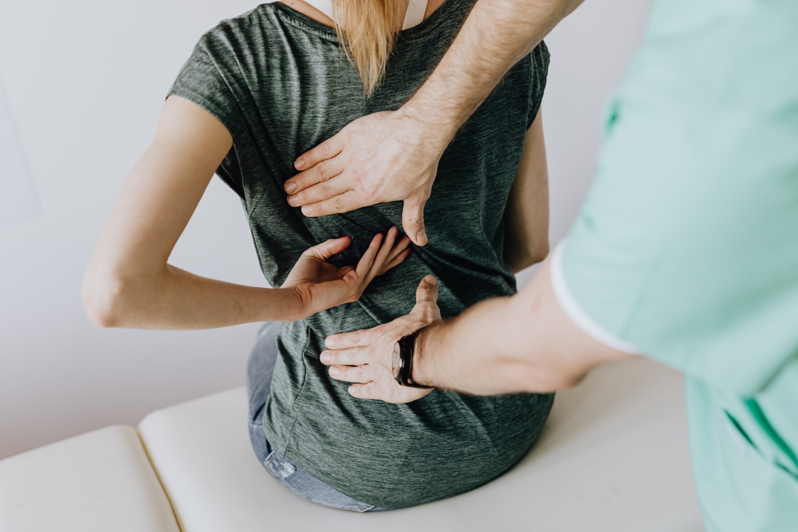 A woman sitting on an examining table showing a doctor an area of her back with her hand.