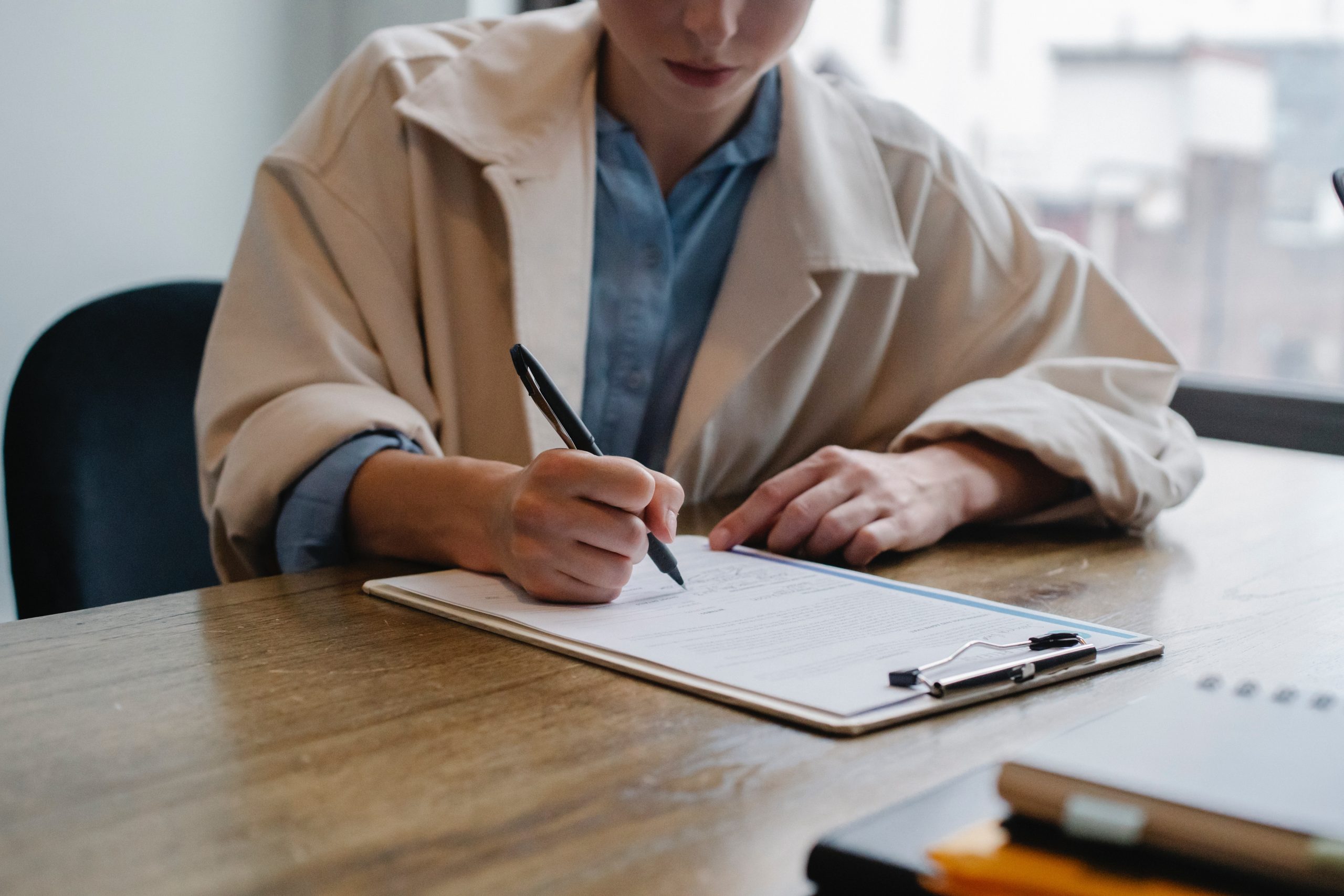 A woman in a trench coat sits at a table and fills out an application form.
